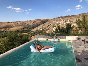 Une femme flotte sur un radeau gonflable dans une piscine avec une vue panoramique de collines ondulantes et d'arbres sous un ciel bleu clair.