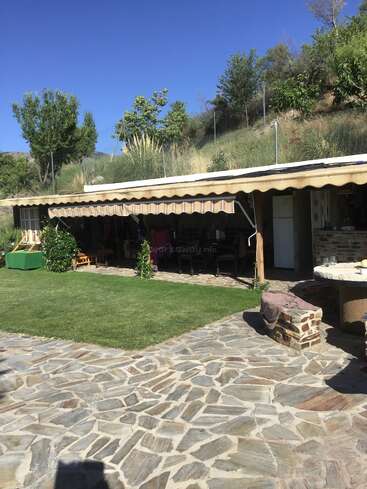 L'image représente un cadre extérieur serein, avec une maison dotée d'un patio, un sentier en pierre, une zone herbeuse et une colline avec des arbres et des buissons, sous un ciel bleu limpide.