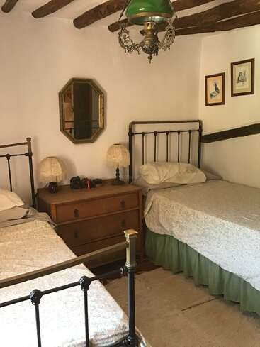 L'image représente une chambre à coucher confortable avec deux lits, une commode et un miroir, avec des murs blancs, des poutres en bois et un lustre, dégageant une atmosphère chaleureuse et accueillante.
