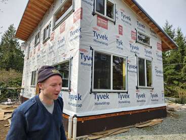 Un homme coiffé d'un bonnet se tient devant une maison en construction, recouverte d'une membrane Tyvek HomeWrap. La scène se déroule en plein air, au milieu des arbres.