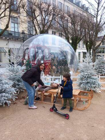 Eine Frau mit zwei Kindern posiert neben einer Schlittenbank, umgeben von verschneiten Bäumen, einem Roller und einer festlichen Weihnachtskugel in einer geschmückten Außenanlage.