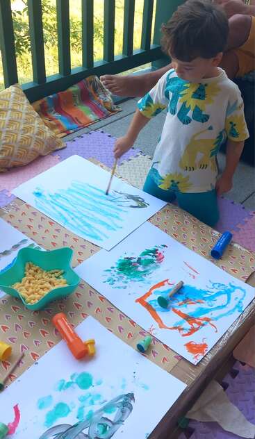 Ein kleines Kind malt kreative, farbenfrohe Bilder auf weißem Papier, umgeben von Malutensilien und Snacks, auf einer Matte im Freien sitzend, und genießt einen spielerischen, künstlerischen Moment.