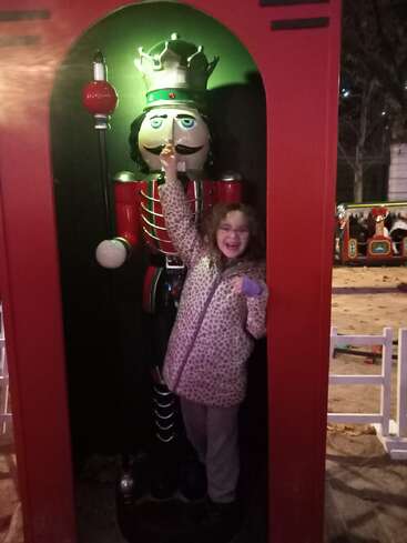 Ein fröhliches Mädchen steht neben einer großen Nussknacker-Statue in festlicher Umgebung, lächelt strahlend, trägt eine gepunktete Jacke und berührt spielerisch die Nase des Nussknackers. Nächtliche Szene.