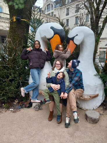 Eine fröhliche Familie posiert spielerisch vor einer großen Schwanenskulptur. Umgeben von Bäumen und städtischen Gebäuden lächeln alle und genießen gemeinsam einen lustigen Moment im Freien.