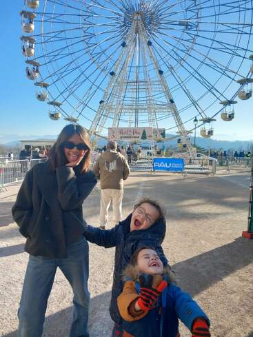 Eine Frau und zwei fröhliche Kinder posieren an einem sonnigen Tag vor einem großen Riesenrad, lachen und genießen eine festliche Veranstaltung im Freien.