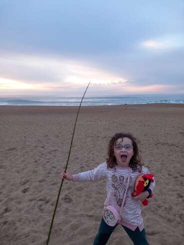 Ein fröhliches Kind steht bei Sonnenuntergang an einem Sandstrand, hält einen langen Stock und ein Plüschtier in der Hand und lächelt breit unter einem blau-rosa Himmel.