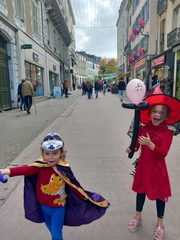 Zwei Kinder in bunten Kostümen, eines als Hexe mit einem Luftballon, das andere mit Maske und Umhang, feiern auf einer belebten europäischen Straße voller Fußgänger.