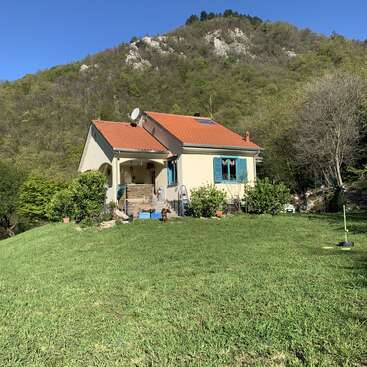 L'image représente une charmante maison de plain-pied au toit orange vif, nichée dans une cour verdoyante, entourée d'arbres et d'une majestueuse montagne en toile de fond.