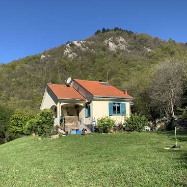 The image depicts a charming single-story house with a vibrant orange roof, nestled in a lush green yard, surrounded by trees and a majestic mountainous backdrop.