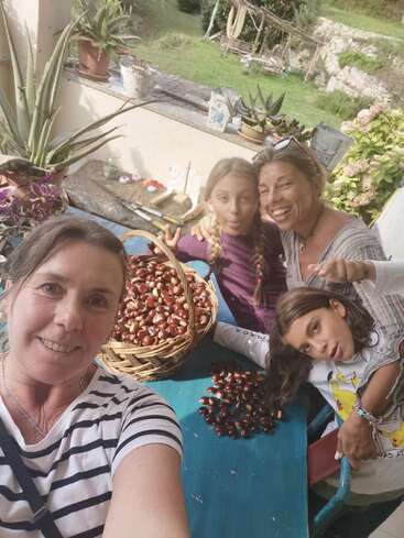 Vier Menschen lächeln und posieren für ein Selfie um einen blauen Tisch mit Kastanien. Sie genießen einen gemütlichen, sonnenbeschienenen Moment auf einer Terrasse mit üppigem Grün draußen.