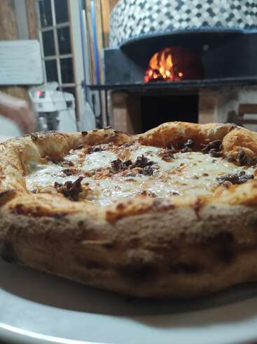 Una deliciosa pizza de masa gruesa cubierta con queso fundido y carne reposa en un plato, con un horno de leña ardiendo cálidamente al fondo. Ambiente rústico.