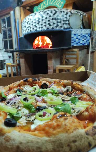 Una pizza recién horneada con aceitunas, pimientos y albahaca se encuentra en primer plano. Detrás, un horno tradicional de leña brilla cálidamente en una acogedora pizzería.