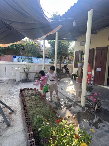 Zwei Kinder stehen in einem sonnigen Garten und gießen ein kleines, von Ziegelsteinen umgebenes Beet, im Hintergrund sind bunte Blumen, Pflanzen und ein gemütliches Häuschen zu sehen.