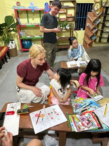 Eine Gruppe von Leuten, darunter auch Kinder, sitzt mit Malbüchern und Malutensilien an kleinen Tischen und genießt kreative Aktivitäten in einem gemütlichen, mit Büchern gefüllten Raum.