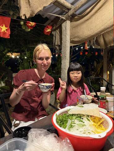 Eine Frau und ein junges Mädchen genießen im Freien eine Mahlzeit, umgeben von vietnamesischen Flaggen und frischem Gemüse. Das Mädchen macht ein Friedenszeichen und lächelt herzlich.