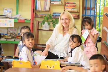 Eine lächelnde Lehrerin sitzt, umgeben von kleinen Kindern, in einem hellen Klassenzimmer voller Bücher, bunter Stühle und fröhlicher Dekorationen, was eine warme und einladende Lernatmosphäre schafft.