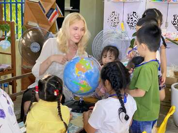 Eine blonde Frau unterrichtet eine Gruppe kleiner Kinder in Geografie mithilfe eines Globus in einem bunten, fröhlichen Klassenzimmer mit Büchern, Ventilatoren und Dekorationen.