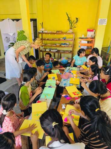 Eine Gruppe von Kindern sitzt an Tischen und bastelt mit buntem Papier. Eine Lehrerin beugt sich vor, hilft ihnen und unterhält sich mit ihnen in einem leuchtend gelben Klassenzimmer.