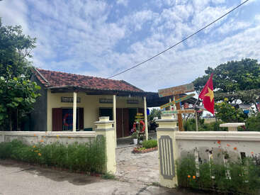 Hinter einem niedrigen Zaun steht ein kleines Haus mit rotem Ziegeldach. Eine vietnamesische Flagge weht im Wind, üppiges Grün und Wegweiser runden das friedliche Bild ab.