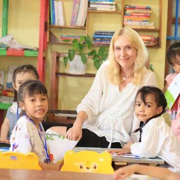A smiling teacher sits surrounded by young children in a bright classroom filled with books, colorful chairs, and lively decorations, creating a warm and welcoming learning environment.