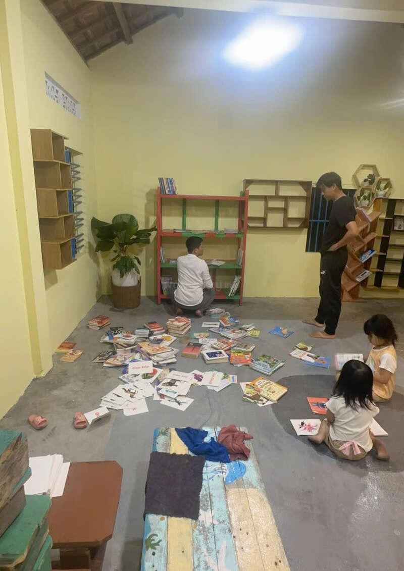 Vier Kinder und ein Erwachsener sortieren und lesen viele verstreute Bücher in einem gemütlichen Raum mit Bücherregalen, Pflanzen und einer entspannten, einladenden Atmosphäre.