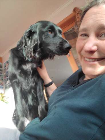 Une personne souriante pose pour un selfie avec un chien noir amical. Le chien est assis tout près, montrant de l'affection. La lumière du soleil illumine l'intérieur douillet.