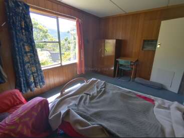 Une chambre à coucher douillette, lambrissée, avec une grande fenêtre qui laisse entrevoir la verdure à l'extérieur. Un lit non fait, une armoire, un bureau avec une chaise et des rideaux colorés complètent cet espace simple et rustique.