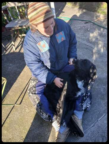 A woman wearing a striped beanie and blue jacket sits outside on the ground, lovingly holding a relaxed black dog in her lap, enjoying the sunshine.