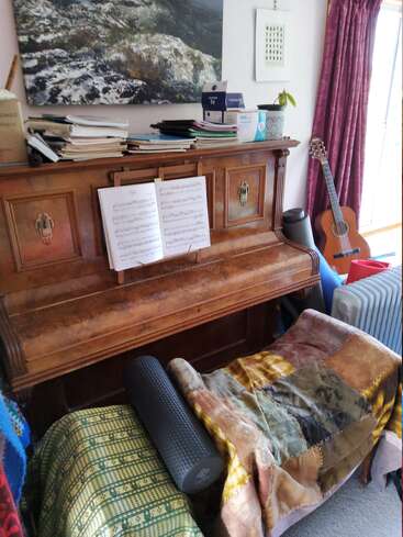 A cozy room features an old upright piano with sheet music, books, a potted plant, a guitar, blankets, a foam roller, and deep red curtains.