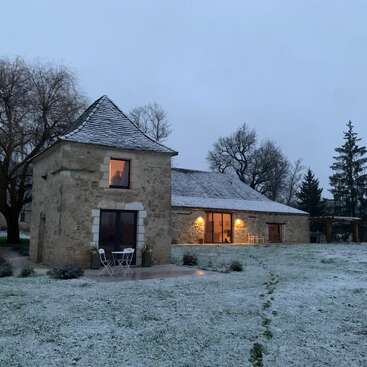 Das Bild zeigt ein ruhiges Steinhaus mit einem schneebedeckten Dach, das in einem frostigen Feld steht, umgeben von Bäumen und beleuchtet von warmen Lichtern, die von den Fenstern und Türen ausgehen.