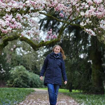Une femme vêtue d'un manteau sombre et d'une écharpe marche sur une allée du parc recouverte de pétales tombés, sous des branches de magnolia en fleurs, avec une végétation luxuriante en arrière-plan.