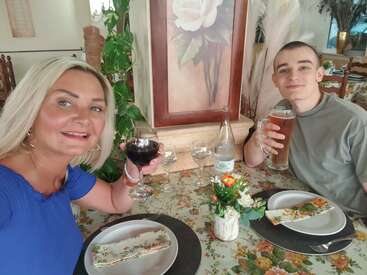 Das Bild zeigt eine Frau und einen Mann, die an einem Tisch sitzen, jeder hält ein Glas Wein oder Bier in der Hand, vor ihnen stehen Teller mit Essen.