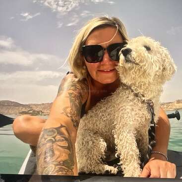 Eine Frau mit blonden Haaren, Sonnenbrille und Tattoos lächelt, während sie ein Selfie auf einem Boot mit ihrem gelockten weißen Hund macht. Ein schöner sonniger Tag im Freien.