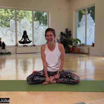 Una mujer se sienta con las piernas cruzadas sobre una esterilla de yoga, sonriendo felizmente en un luminoso estudio con grandes ventanales, plantas y artísticos diseños de lotos y chakras en las paredes.