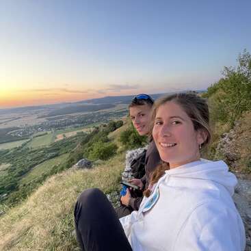 Un couple souriant est assis sur une colline herbeuse au coucher du soleil, surplombant une vallée pittoresque avec des champs, des villages lointains et des montagnes sous un ciel clair. Un moment paisible.