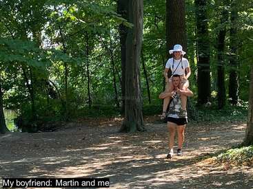 Una pareja disfruta de un paseo por un bosque verde e iluminado por el sol. El hombre lleva a su novia a hombros mientras ambos sonríen, rodeados de altos árboles y naturaleza.