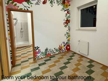 Esta imagen muestra una habitación muy luminosa con coloridos murales florales, baldosas estampadas, una ventana, un radiador y una puerta abierta que conduce a una ducha.