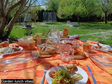 Un vibrante jardín con una mesa puesta para comer. Platos coloridos, ensaladas, bebidas y cristalería crean un ambiente alegre y relajado para comer al aire libre bajo los árboles.
