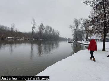 Una persona con abrigo rojo camina por la orilla de un río nevado, rodeada de árboles desnudos y aguas tranquilas, disfrutando de una apacible escena invernal, a pocos minutos de distancia.