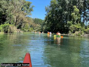 Un grupo de personas navega tranquilamente en kayak por un río claro y verde rodeado de frondosos árboles en un día soleado. El pie de foto dice: "Szigetköz - nuestro hogar".
