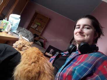 A person in a colorful plaid shirt with headphones around their neck smiles, sitting indoors with an orange cat resting contentedly on their lap beside a desk.