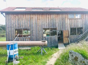 Un edificio rústico de madera con techo de tejas rojas se alza en una zona cubierta de hierba. Se pueden ver grandes ventanas, una puerta abierta, un barril azul y un camino.