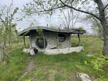 Una encantadora y rústica cabaña de piedra con techo de paja y ventanas redondas se encuentra enclavada en una zona boscosa, frondosa y verde, que evoca una atmósfera de cuento de hadas.