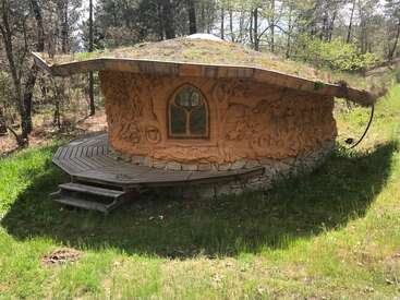 Esta caprichosa casa redonda tiene un techo cubierto de madera y musgo, paredes talladas ornamentadas, cimientos de piedra y un pequeño porche, y está enclavada en un soleado claro del bosque.