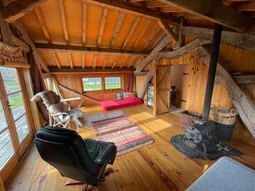Esta acogedora habitación rústica cuenta con vigas de madera, techo inclinado, estufa de leña, silla negra, diván, alfombras tejidas, mecedora y luz natural brillante.