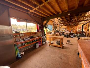 Se trata de un taller espacioso y rústico con techos y vigas de madera, estanterías repletas, bancos de trabajo, herramientas colgadas, iluminación natural y un ambiente organizado y acogedor.