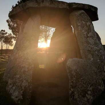 Un dolmen, constitué de grosses pierres anciennes, se dresse dans un champ verdoyant. Le soleil filtre à travers la structure, créant une scène mystique au milieu des arbres.