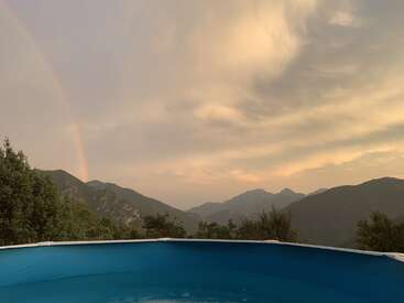 The image depicts a serene pool scene with a majestic mountain range in the background, set against a cloudy sky with a vibrant rainbow.