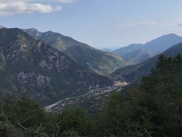 The image depicts a serene mountainous landscape with a town nestled at its base, surrounded by lush greenery and set against a clear blue sky.