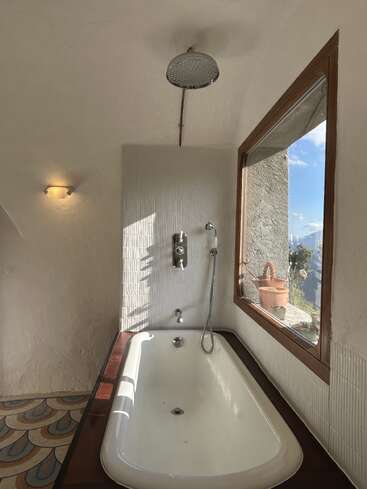 A cozy white bathroom features a bathtub beneath a rain shower, warm wall light, patterned floor, large window with sunlight, and a scenic outdoor view.